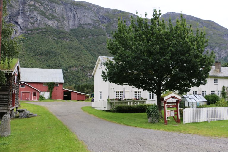 Rød låve, stabbur og et hvitt våningshus på gårdstun med stort, grønt tre. Skogkledd fjellside i bakgrunnen.