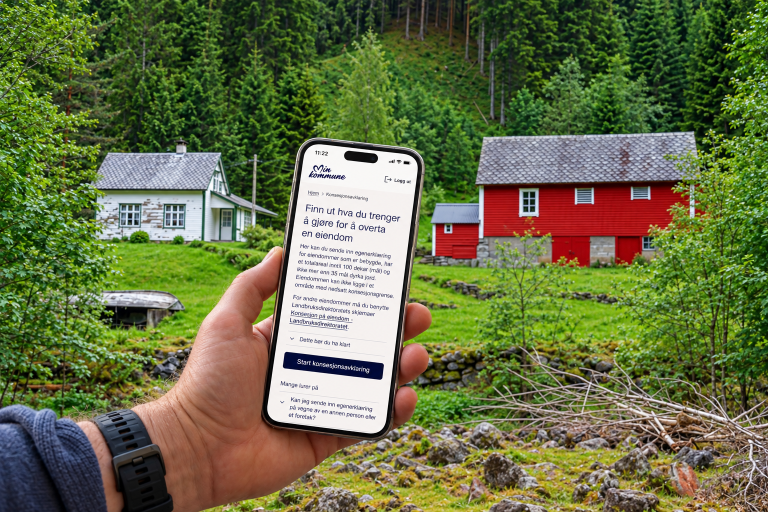 Et hvitt og et rødt hus i skogen, med en hånd i forgrunnen som holder en mobiltelefon med visning av nettsiden "Min kommune"